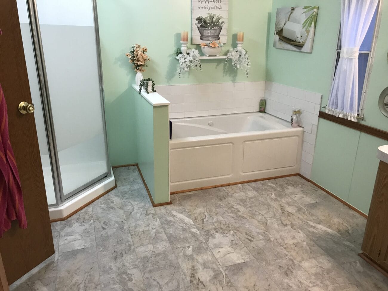 Bathroom with a corner shower, a large bathtub, mint green walls, tiled floor, a window with curtains, and decorative shelves holding plants and candles above the tub.