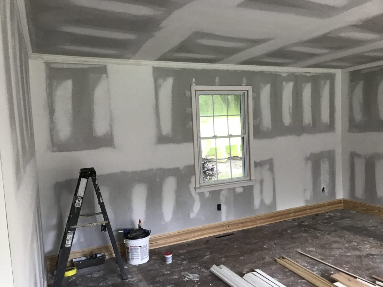 A room under construction with unfinished drywall, a ladder, paint supplies, and scattered wood pieces on the floor. A single window lets in daylight.