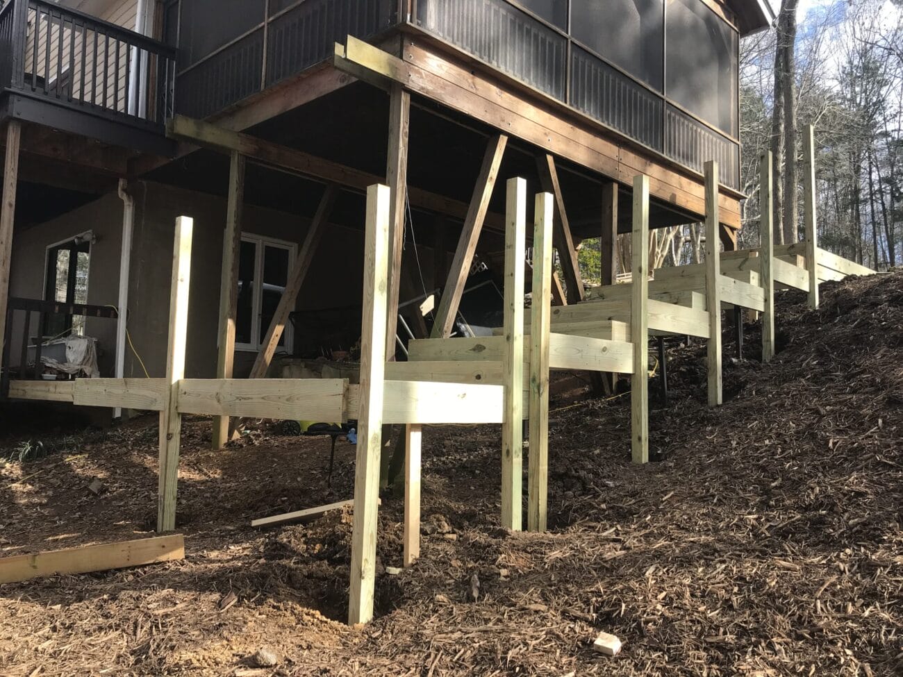 Wooden posts and beams form the framework for a deck or stair structure being built on a sloped yard beside a house.