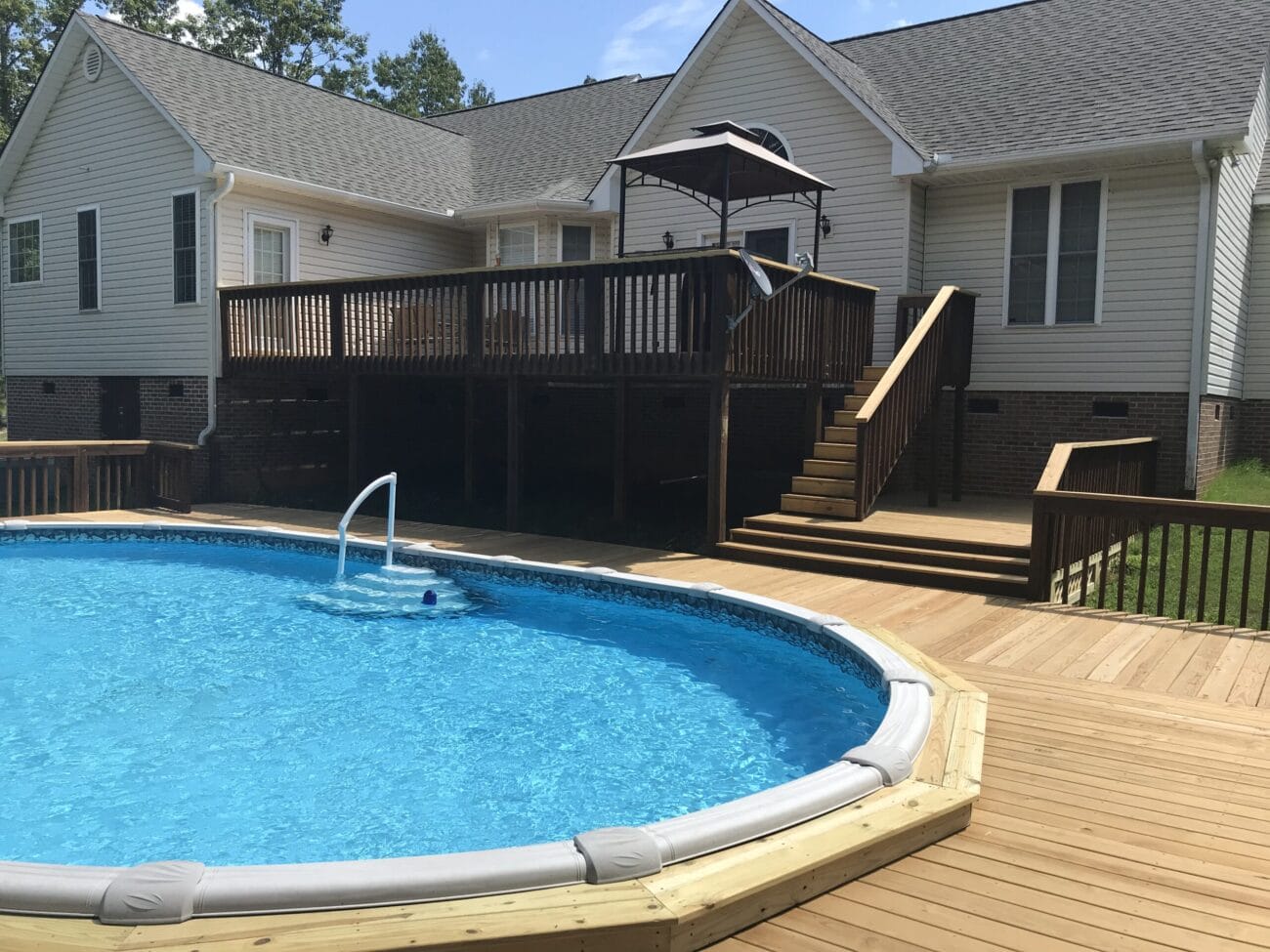 Above-ground pool with clear blue water next to a spacious wooden deck and stairs leading up to a covered patio attached to a light-colored house.