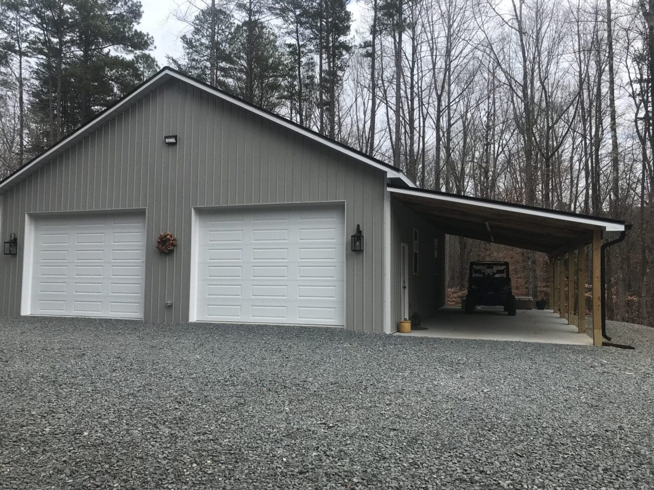 A gray metal building with two white garage doors, a wreath on the left door, and a covered carport on the right, housing an off-road vehicle. Trees are visible in the background.