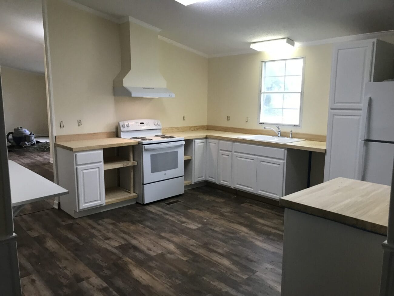 A kitchen with white cabinets, wood countertops, a white stove, range hood, refrigerator, sink under a window, and wood-patterned flooring.