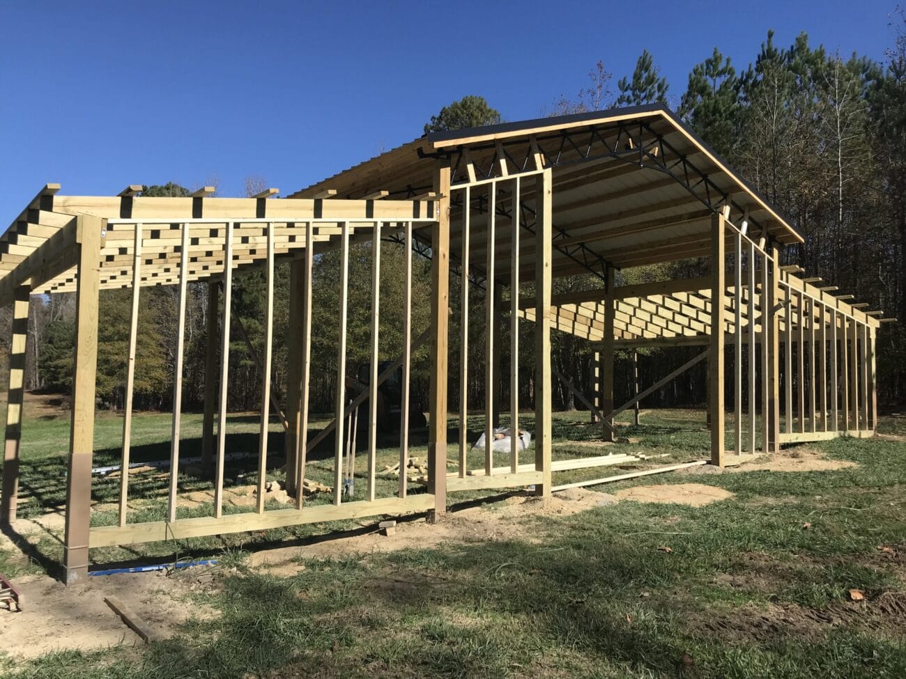 A partially constructed wooden frame structure stands on a grassy area, with trees and a clear blue sky in the background.